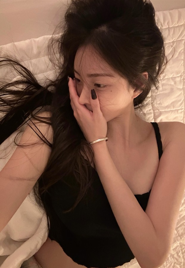 สาวเกาหลีในเสื้อแขนกุดสีดำนั่งบนเตียงสีขาวในมุมห้องพักผ่อนสบายตา