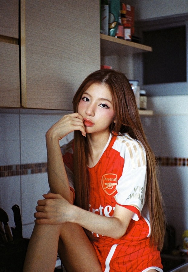 เบลล์ ณัชชา ในเสื้อฟุตบอลสีแดงขาวตัวโคร่งนั่งกอดเข่าบนเคาน์เตอร์ไม้